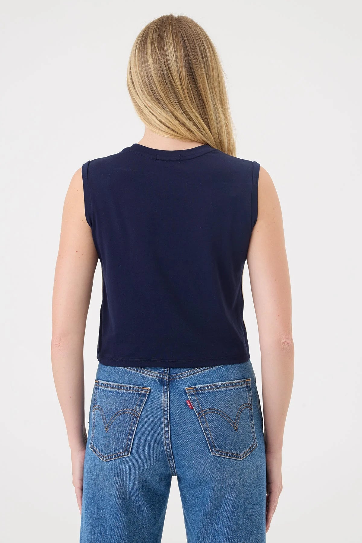 A woman viewed from the back wearing a sleeveless navy blue Goldie top and blue jeans against a plain light gray studio background.