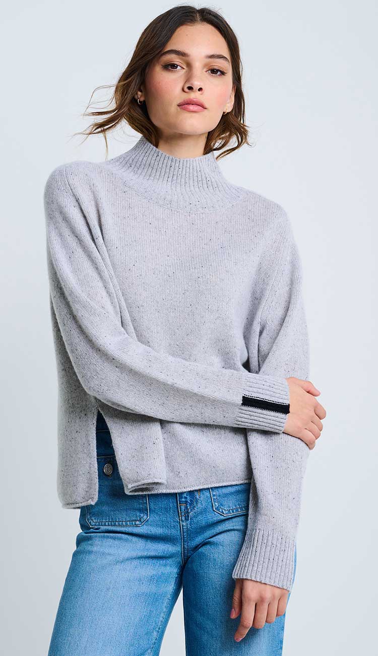 woman wearing the speakeasy sweater in light silver speckled grey with slits on the sides, wearing blue jeans on a light grey background.