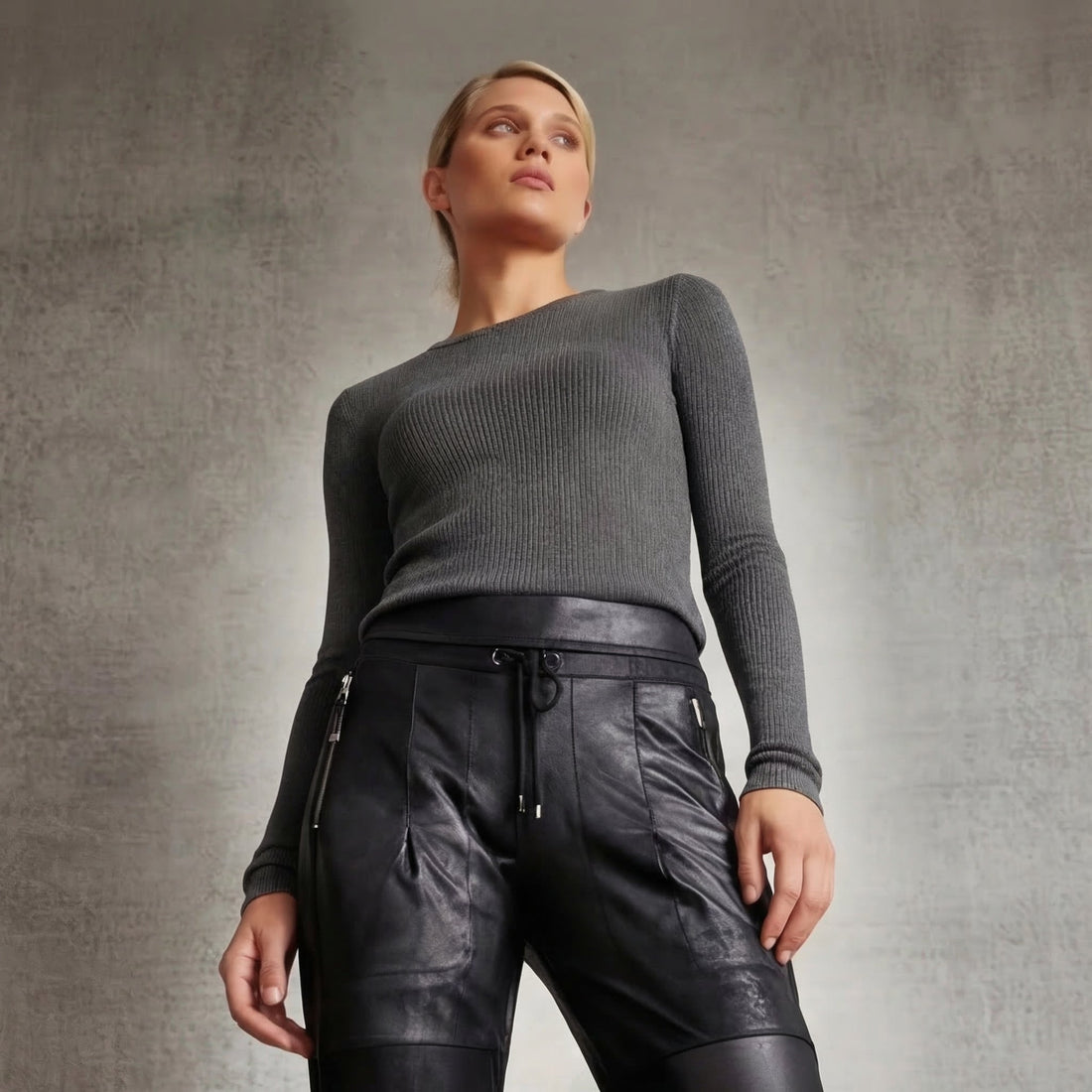 A woman standing in a gray Goldie long sleeve t and vegan leather candy pants in black with a beige background. 