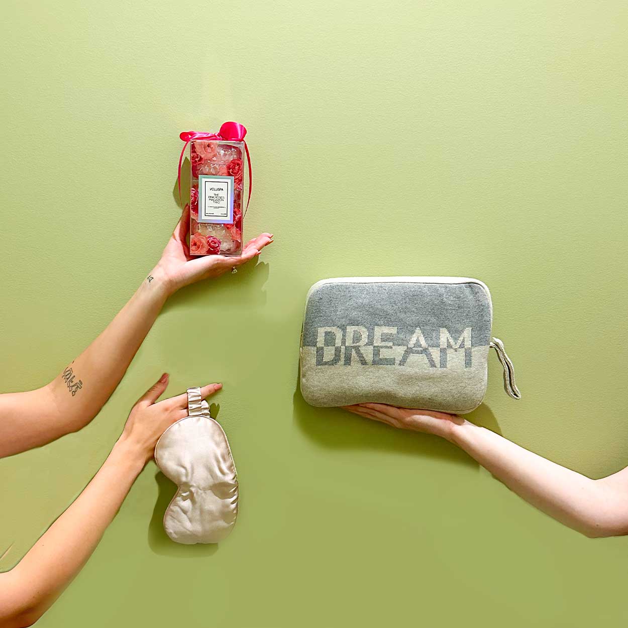 hands and arms of persons holding an silk eye mask, a box of voluspa candles and a pouch with a blanket