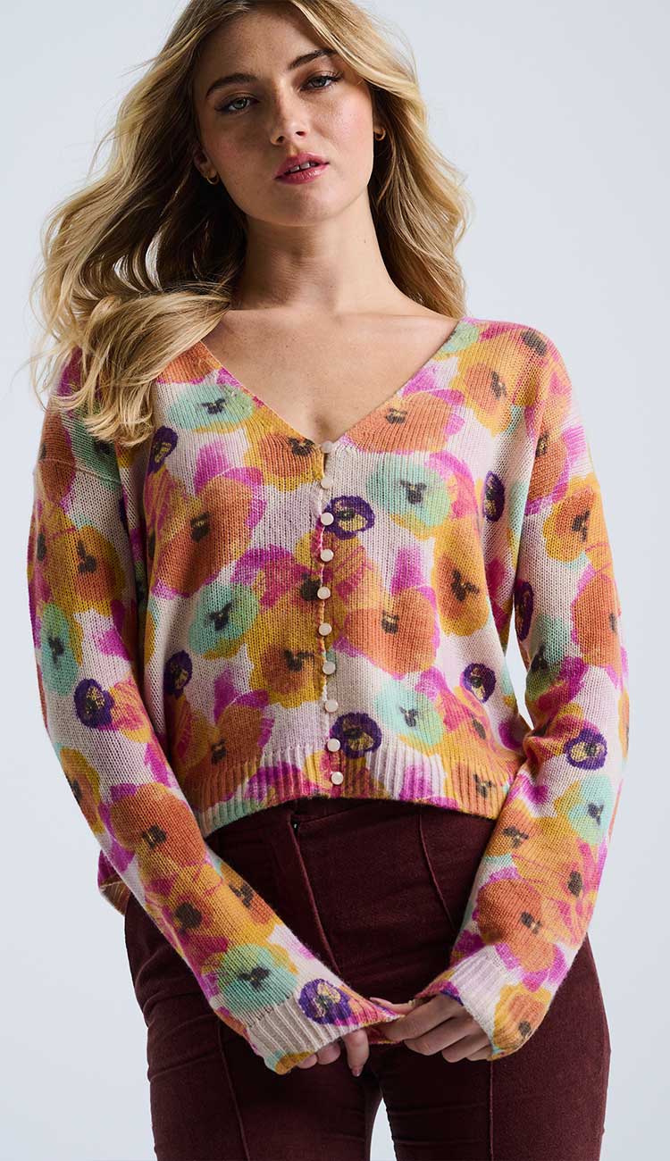 Woman wearing the bloomin' v-neck with little buttons-  sweater with beautiful orange, green a bright pink pansy flowers wearing rust pants on a grey background. 