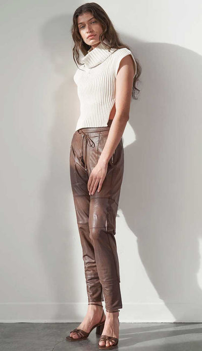 Woman wearing a white sleeveless top and terra brown leather pants against a plain background