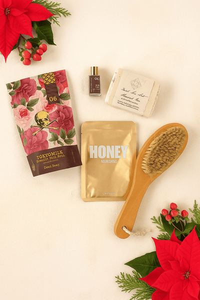Cosmetic items including a body brush, soap, a pouch with bath salts, a gold pouch with a face mask, and a mim bottle of perfume on a light background with poinsettias.