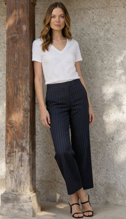 A woman wearing Grace Needle Line Navy Stripe pants with a white Goldie tee, leaning on a wooden column.