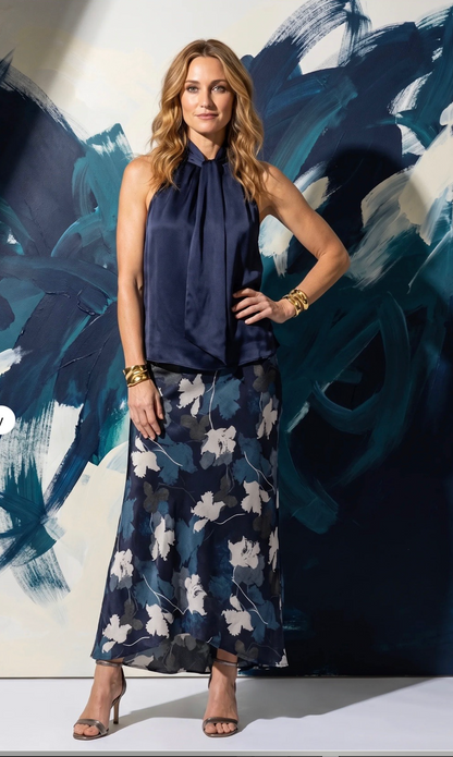 A woman models a sleeveless go silk navy blue halter-neck top paired with a matching navy blue maxi skirt featuring a white floral pattern, posed in front of an abstract blue and white painting.