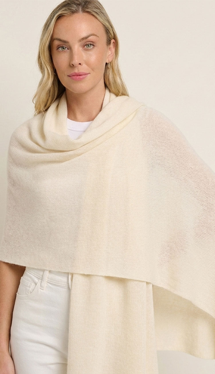 Woman wearing a cream-colored alashan breezy travel wrap over a white outfit on a plain background
