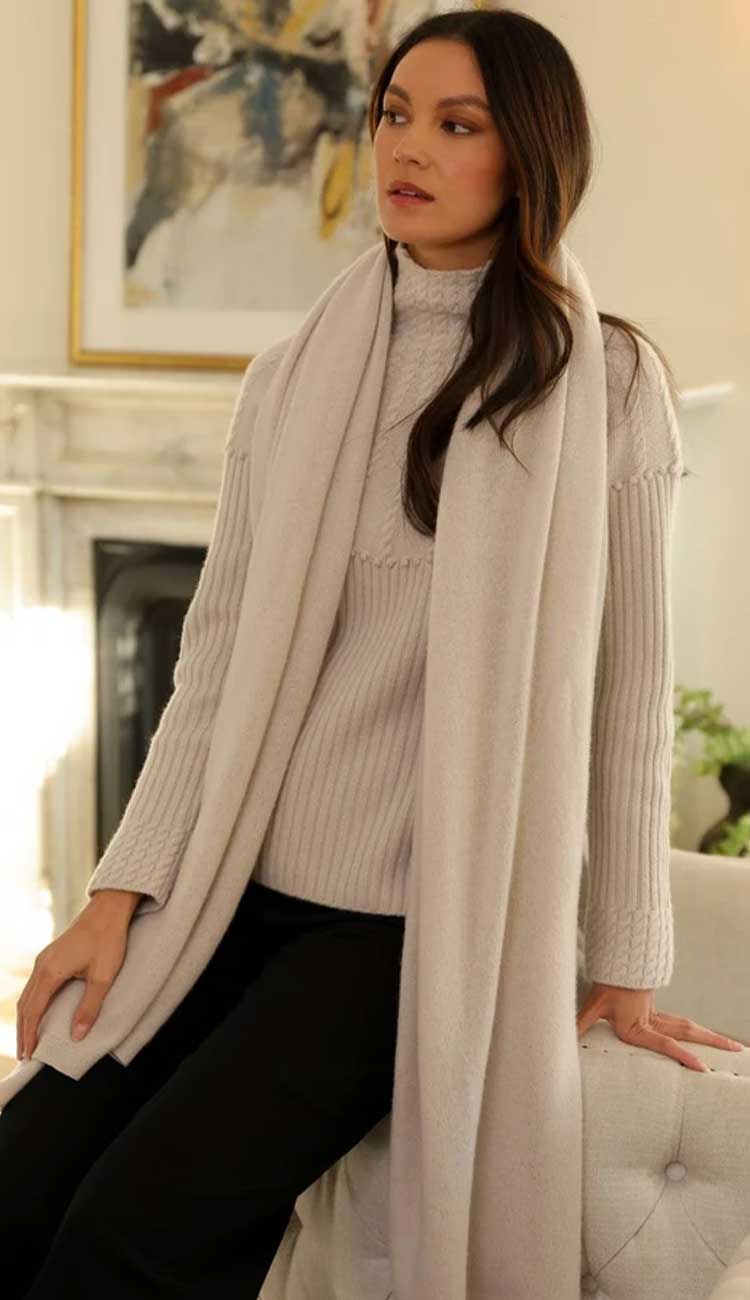 alashan cashmere breezy travel wrap | paula and chlo Champagne model wearing a matching sweater sitting on the side of a couch