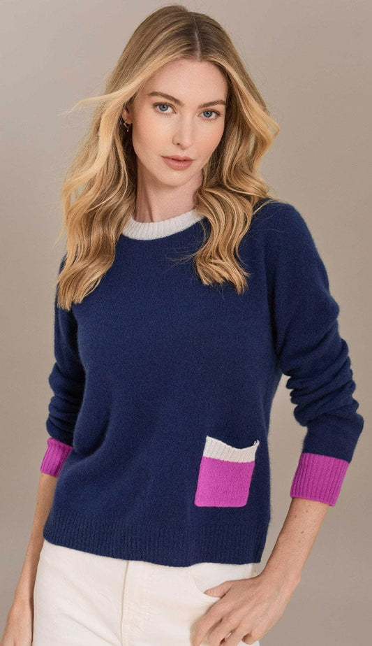 Front view of a woman wearing a navy blue Alashan cashmere crew neck sweater. The sweater features a contrasting white ribbed collar, bright pink ribbed cuffs, and a small patch pocket on the lower left with a pink base and white trim
