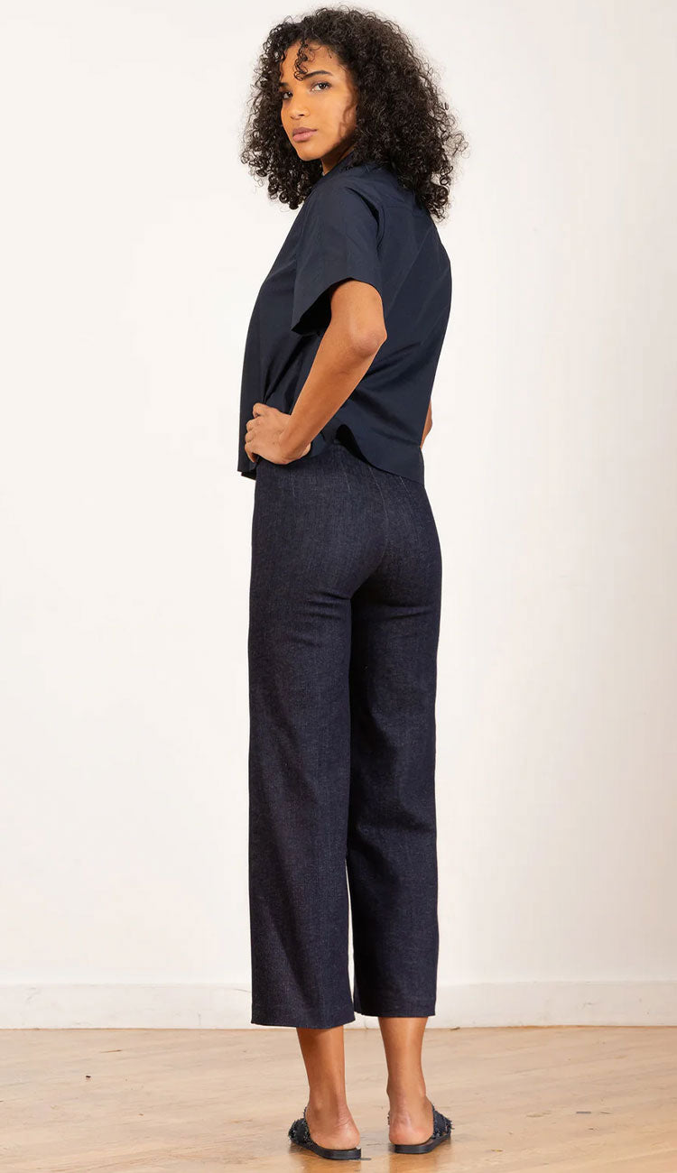 Back view of a woman wearing a dark denim shirt and Alex dark denim pants against a textured wall.