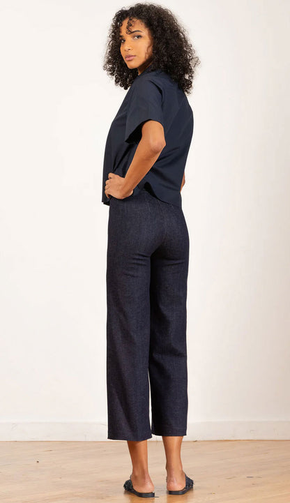 Back view of a woman wearing a dark denim shirt and Alex dark denim pants against a textured wall.