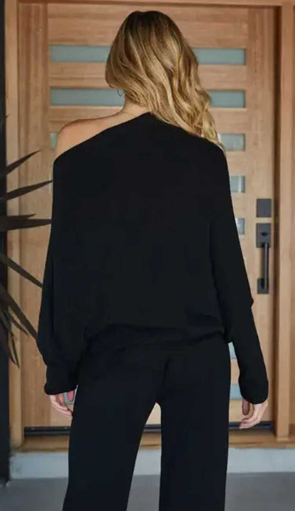 Person wearing a black off-shoulder top and black pants in front of a wooden door.