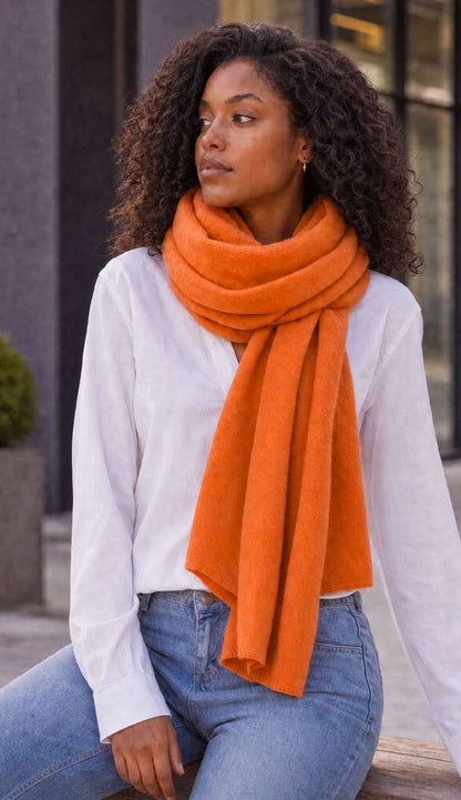 A woman with sits outdoors wearing a White + Warren bright tangerine travel wrap, a white long-sleeve blouse, and light blue jeans.