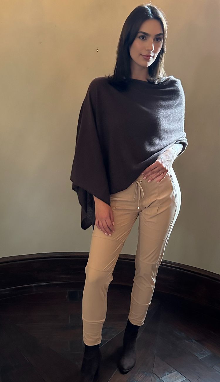Woman wearing a dark gray poncho over a light-colored top and beige pants, standing against a plain wall.