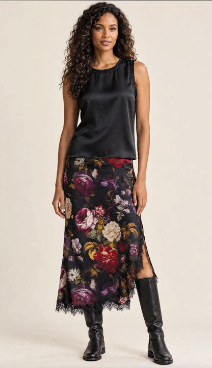 A woman modeling a Go Silk sleeveless black satin top paired with the Go Silk Lacy Floral Print Skirt, a black midi skirt featuring a vibrant red, purple, and white floral print, styled with black leather knee-high boots.