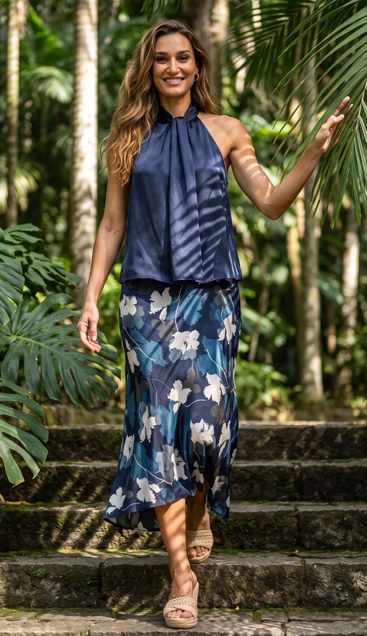A woman wearing a Go Silk navy blue halter top and a matching navy midi skirt with a white floral print, walking down stone steps in a lush tropical garden.
