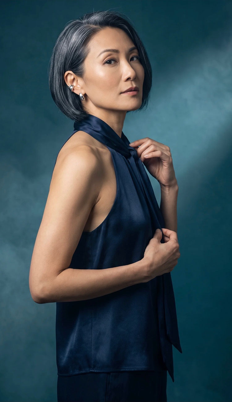 A woman with short gray hair models a dark navy blue satin halter top against a textured dark teal studio background.