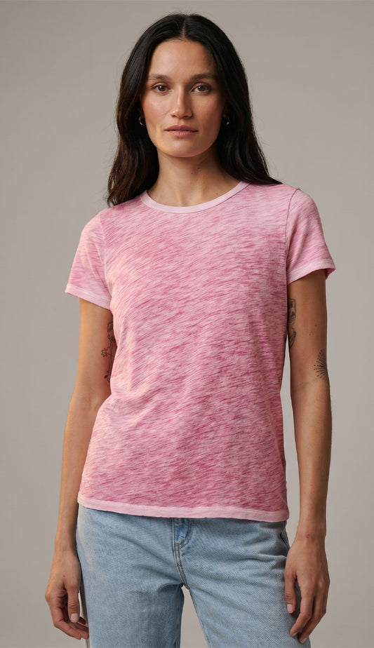 Woman wearing a pink costal sunwash  t-shirt by goldie and blue jeans against a gray background