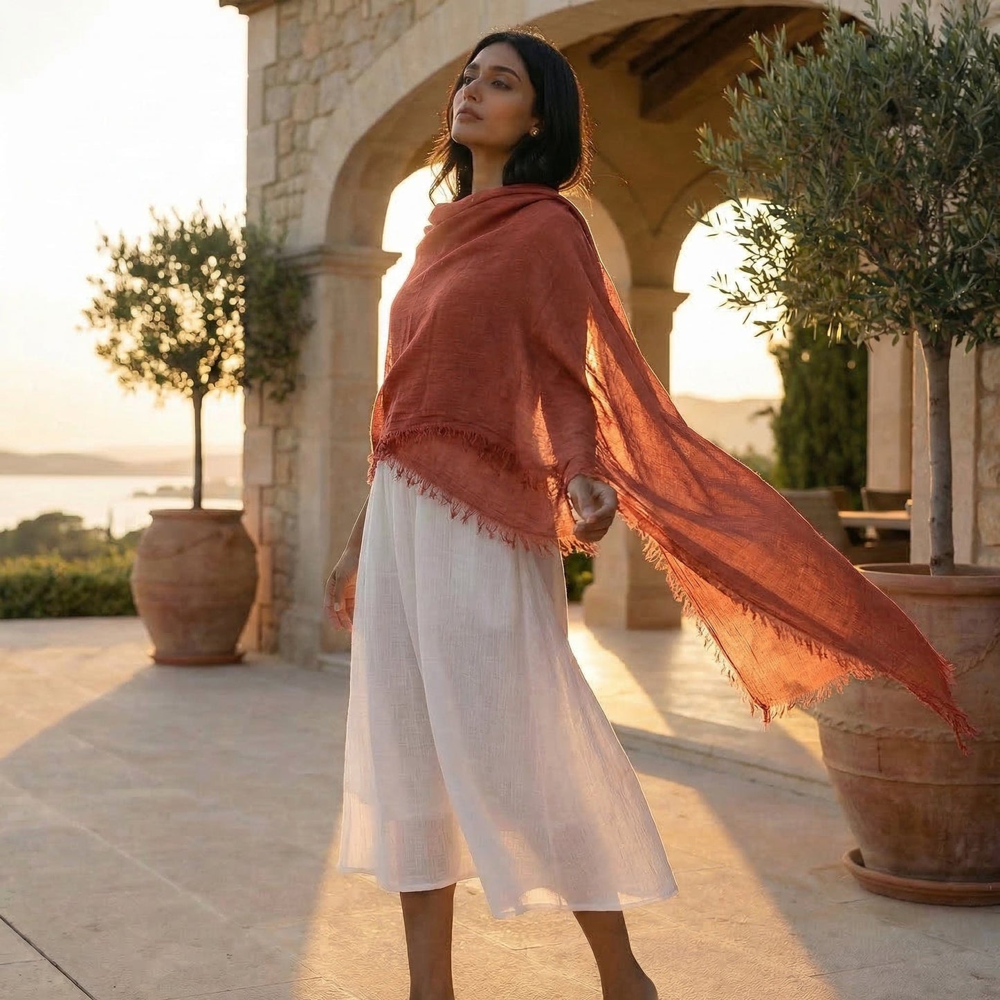 Woman wearing a burnt orange shawl over a white dress in an outdoor setting with stone architecture and potted plants.