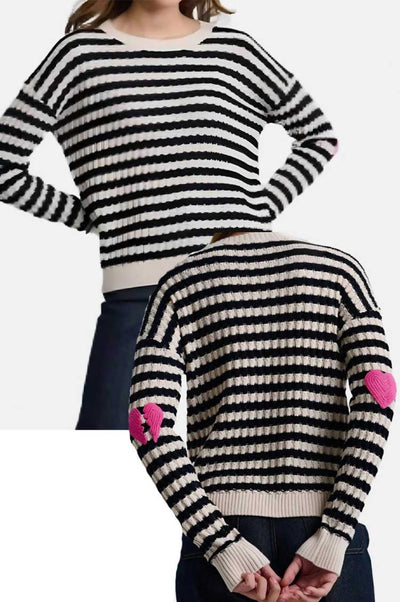Black and white striped sweater with pink heart details on sleeves, worn by a female on a white background.