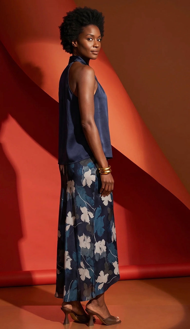A woman modeling a sleeveless navy blue top and a matching long skirt with a white floral pattern, posed against a warm orange and red geometric studio backdrop.