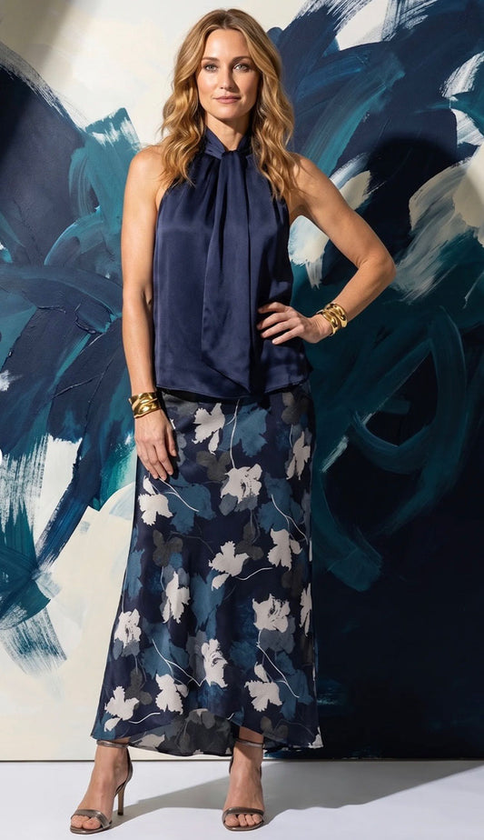A woman models a sleeveless Go Silk navy blue halter-neck top paired with a matching  go silk navy maxi skirt featuring a white floral pattern, standing in front of an abstract blue and white wall.