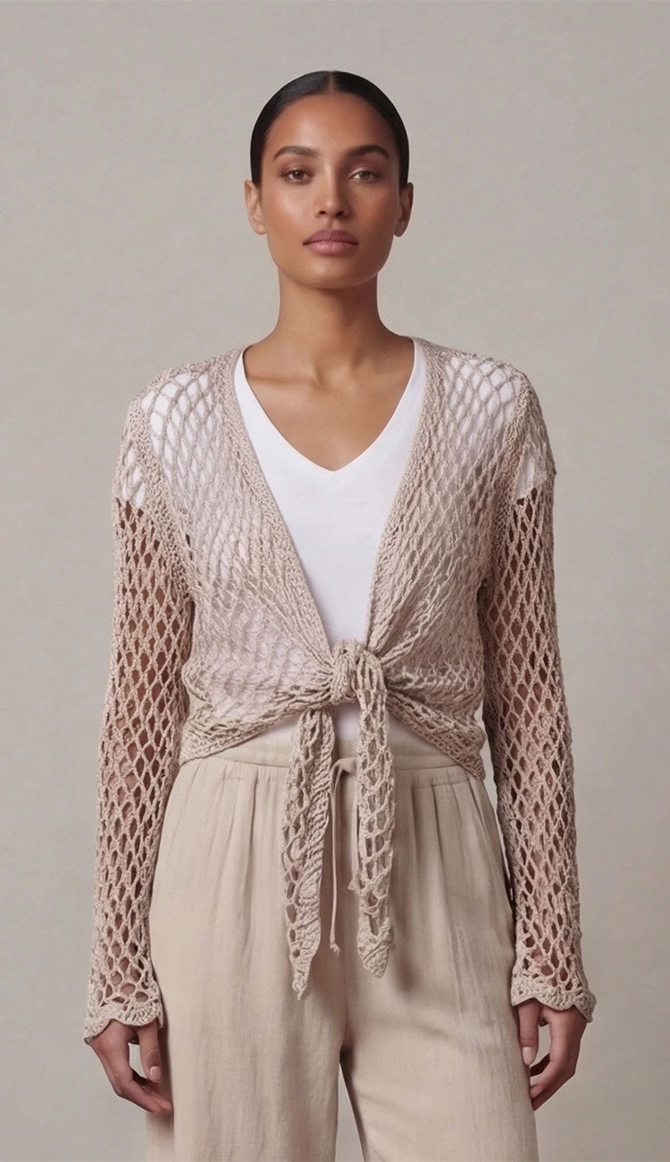 Woman wearing a taupe open crochet top over a white top and beige pants on a plain background