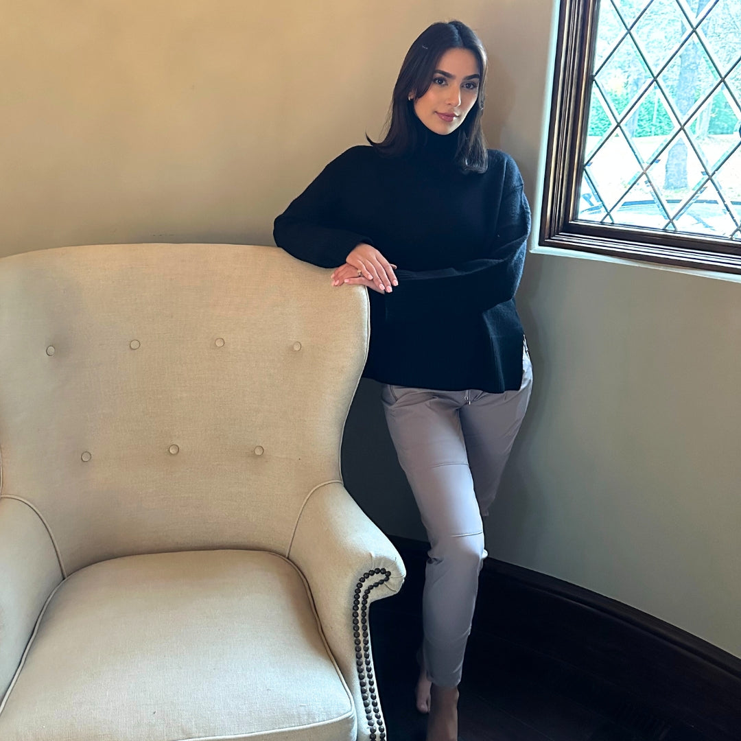 Woman in a black sweater and shadow grey pants standing next to a beige armchair in a room with a window.