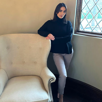 Woman in a black sweater and shadow grey pants standing next to a beige armchair in a room with a window.