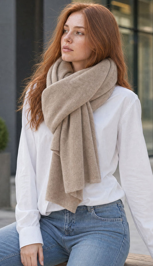 A woman with long red hair sits outdoors wearing the White and Warren Taupe Heather Travel Wrap, a white long-sleeve blouse, and blue jeans.