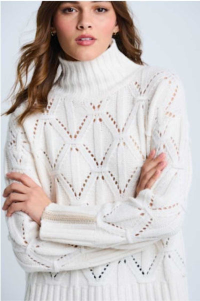 Woman wearing a white detailed knit sweater with a turtleneck against a light background.