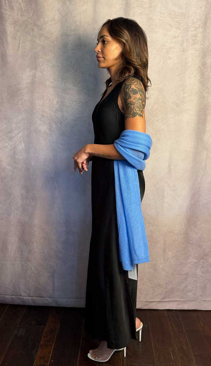 Woman in a black dress with a blue travel wrap draped over her shoulder against a plain background